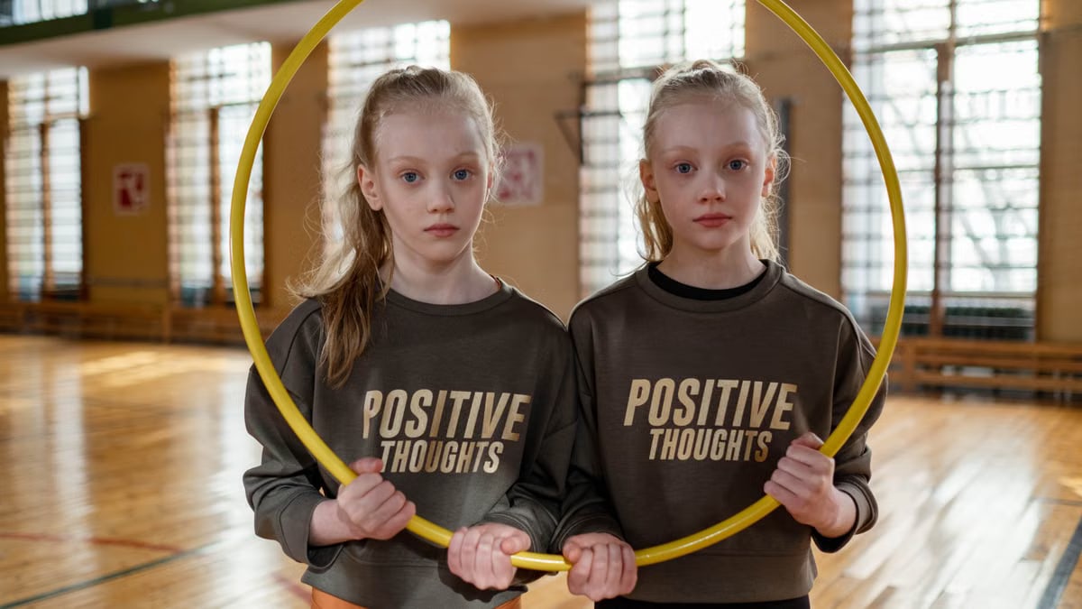 Twin girls in gym holding a hula hoop, wearing 'Positive Thoughts' sweatshirts.